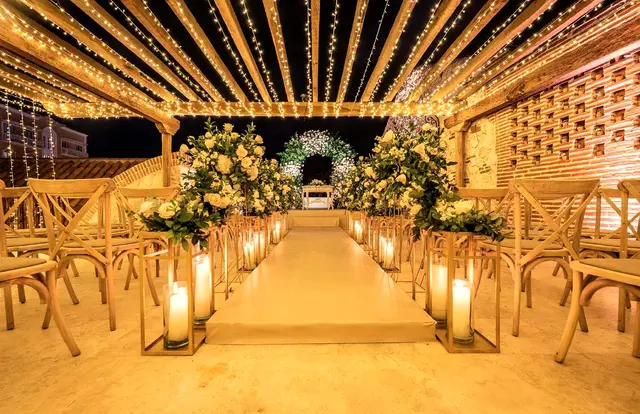 Pasillo de ceremonia iluminado con faroles y un arco de flores bajo un techo de luces