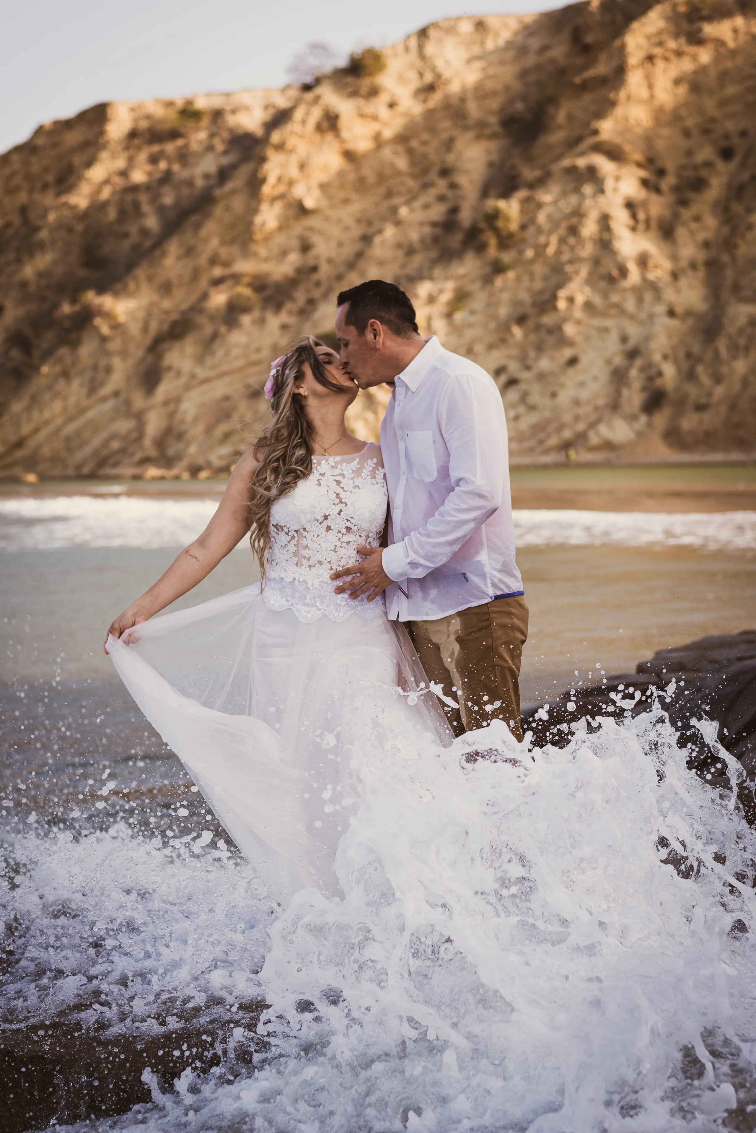 Couple kissing at the shoreline as a wave breaks against the dress