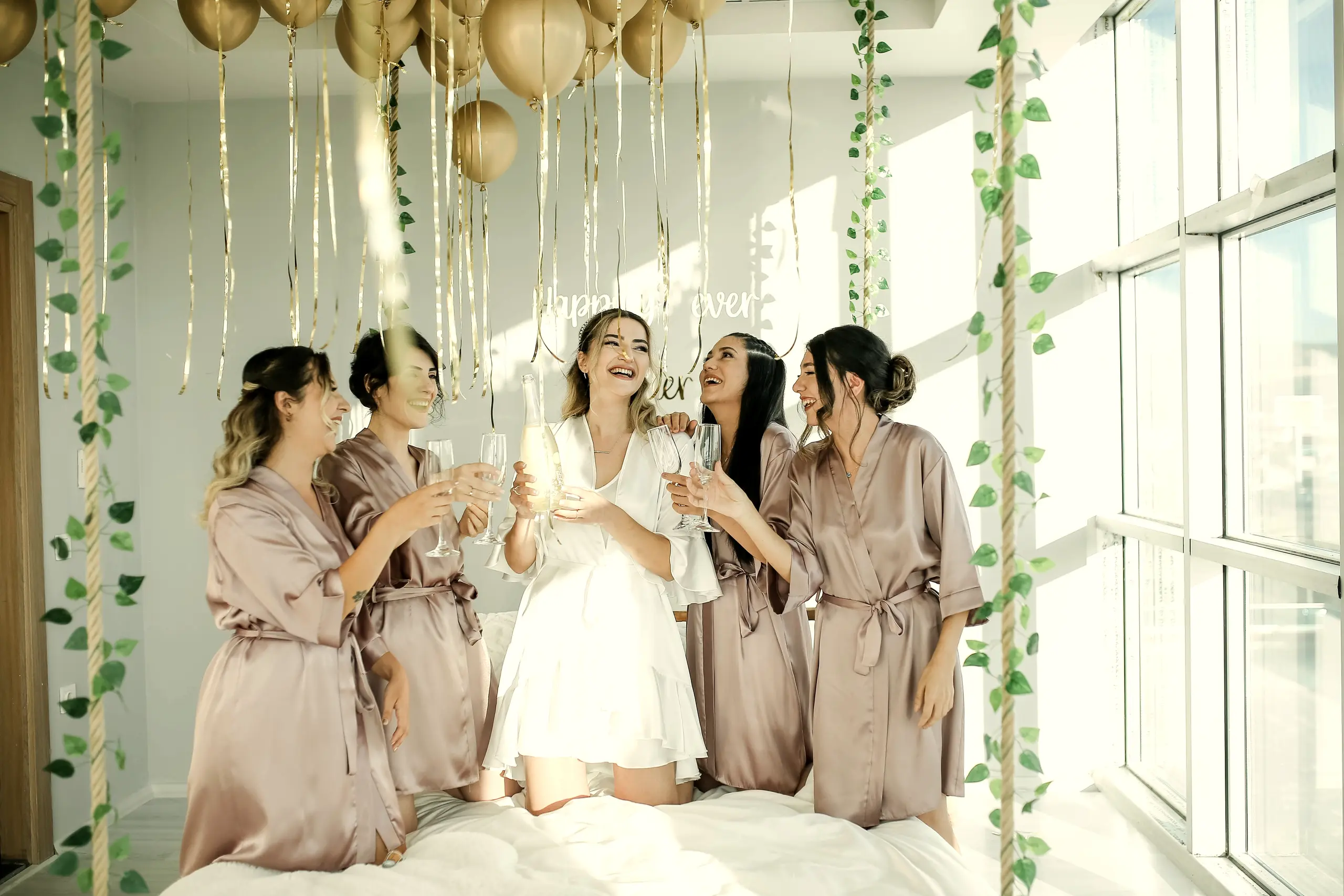 Bride and bridesmaids in champagne satin robes toasting in a room with large windows