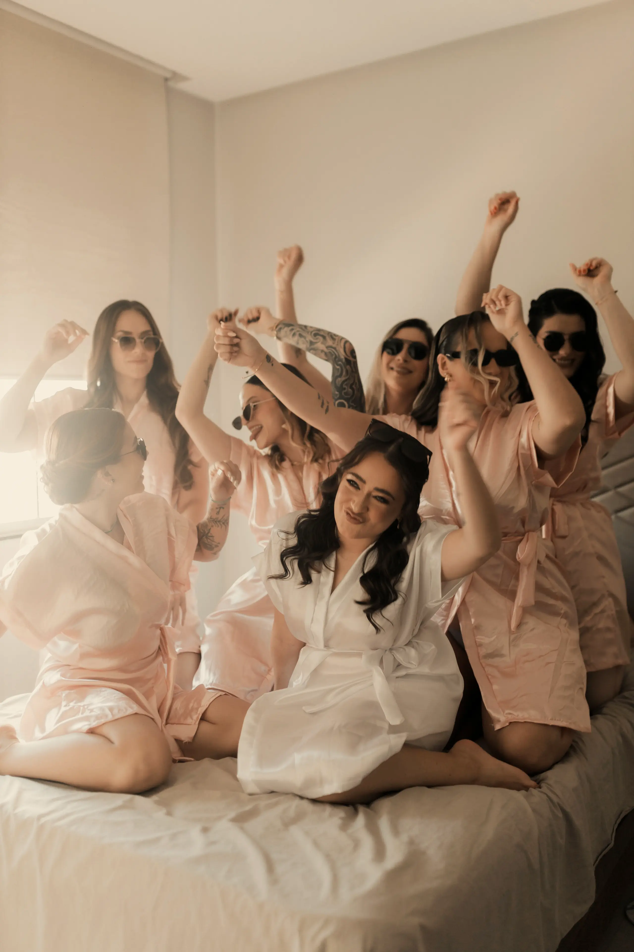 Bride and bridesmaids in pink robes celebrating on a bed with their arms raised