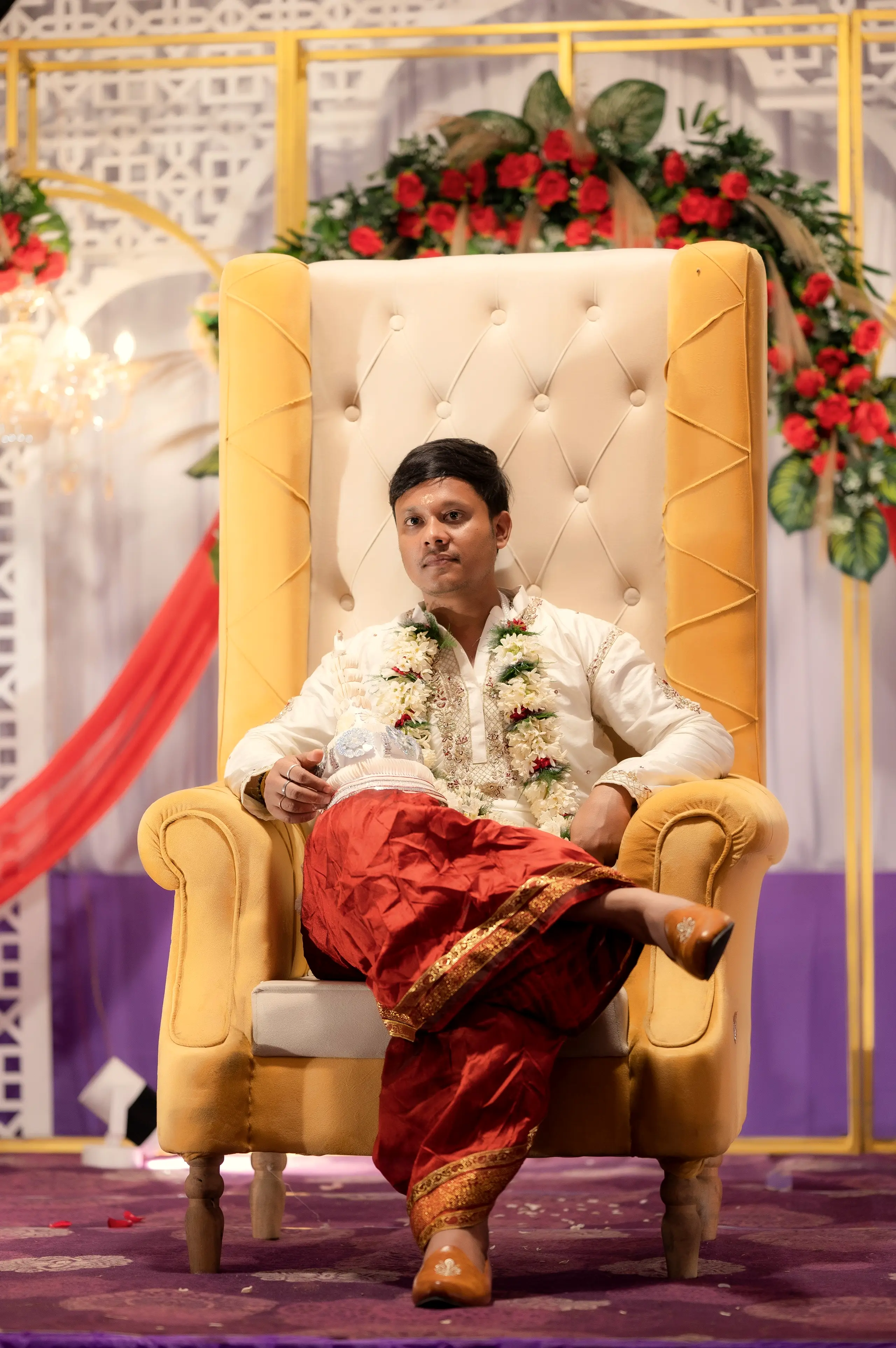 Groom in traditional South Asian attire seated on a golden throne with a flower garland and red fabrics