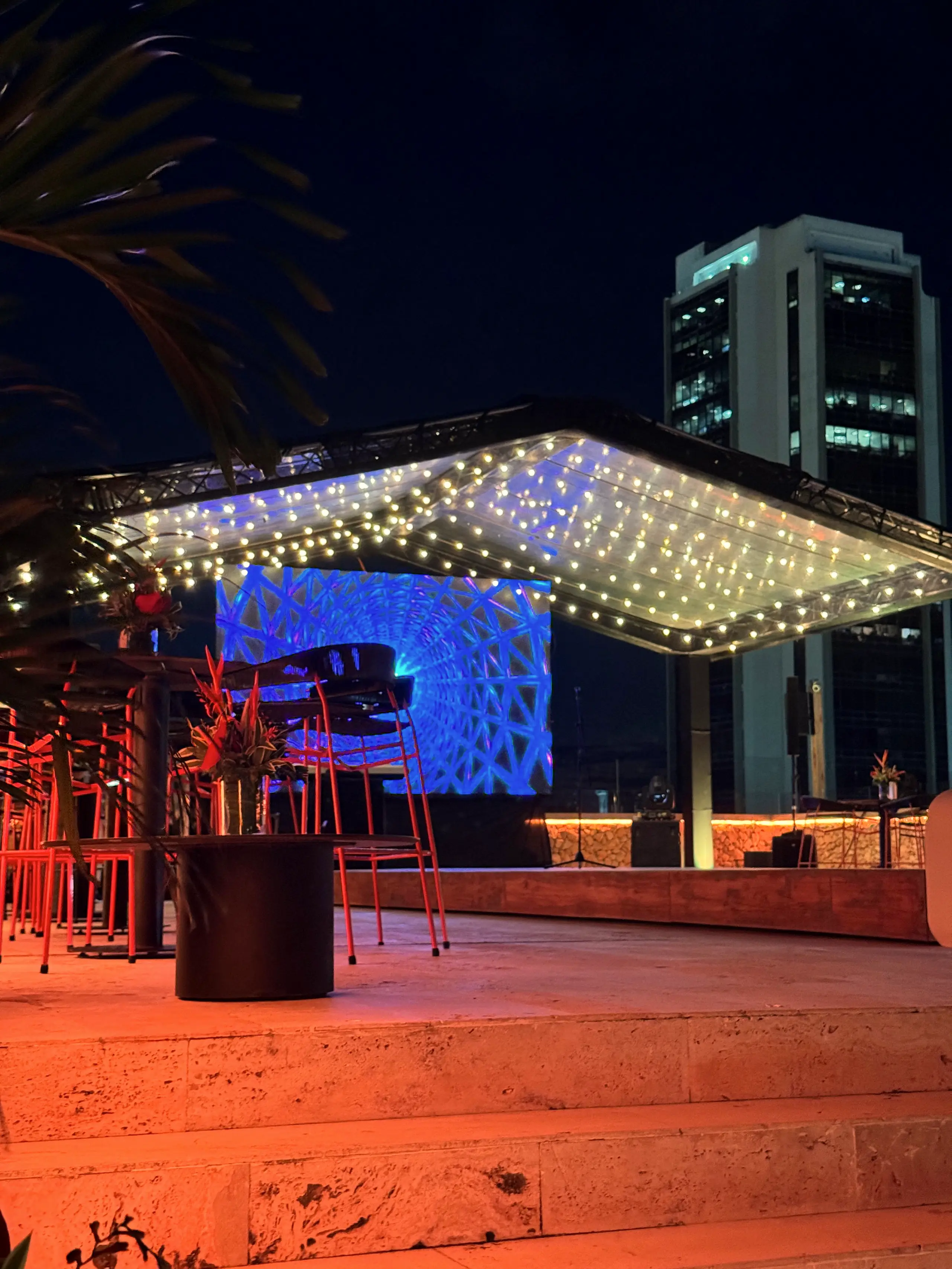 String-light canopy over a blue screen on a terrace with buildings beyond