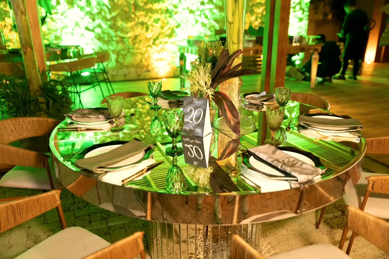 Round mirrored table with tableware and foliage under green light at a night event
