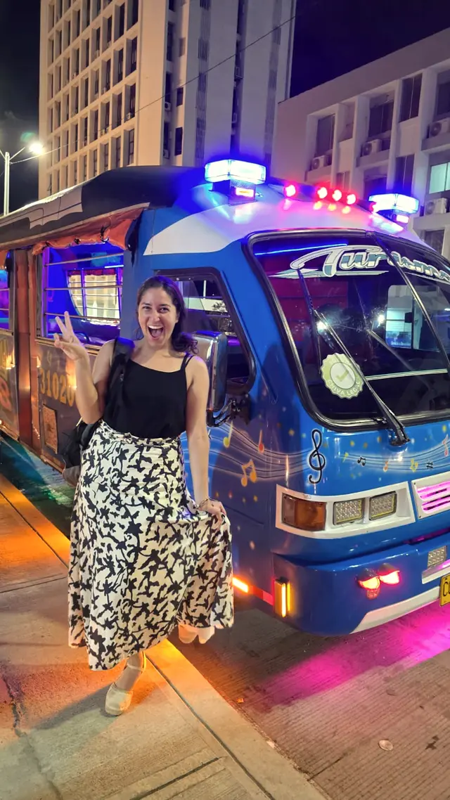 Woman posing beside a blue chiva bus lit with neon lights at night